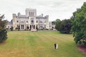 A bride and groom walk hand in hand across a large green lawn in front of a grand historic mansion with ornate architecture, surrounded by trees.