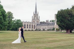 A bride and groom walk hand-in-hand across a large lawn in front of a grand, historic building with a tall spire and arched windows, surrounded by trees.