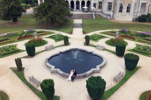 A bride and groom sit together beside a circular fountain in a formal garden with manicured lawns, flowerbeds, hedges, and a historic building in the background.
