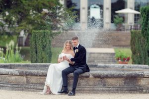 A bride and groom sit together on the edge of a stone fountain, smiling and reading a card. The bride wears a white dress and the groom a black suit. Water sprays behind them in front of a grand building.