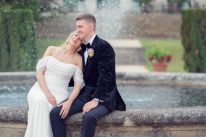A bride in a white off-the-shoulder dress leans her head on the groom’s shoulder as they sit on the edge of a fountain, with water spraying behind them in a garden setting.
