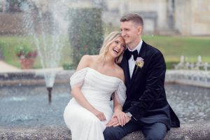A happy bride in a white dress and a groom in a black suit sit together in front of a fountain, smiling and holding hands, with greenery and stone architecture in the background.