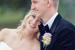A bride leans her head on the groom’s shoulder, smiling warmly. The groom, in a black suit and bow tie, has a buttonhole with a lavender rose. The background is softly blurred with greenery.