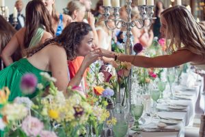 A woman in a green dress leans over a flower-filled table to kiss another woman's hand during a lively indoor celebration with guests standing and mingling in the background.