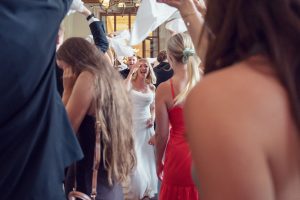 A smiling bride walks through a crowd of guests waving white serviettes in celebration. People in festive attire, including a woman in a red dress, surround her in a bright, joyful indoor setting.