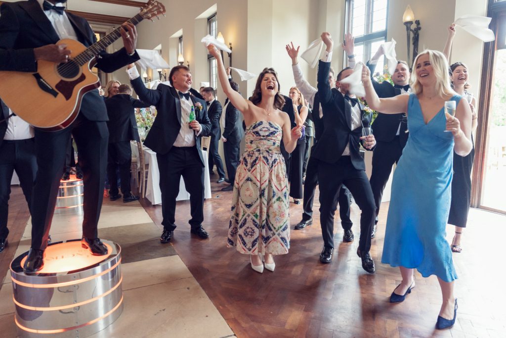 A group of well-dressed people joyfully dance and wave serviettes in the air at a lively indoor celebration, whilst a musician plays guitar on a plinth nearby.
