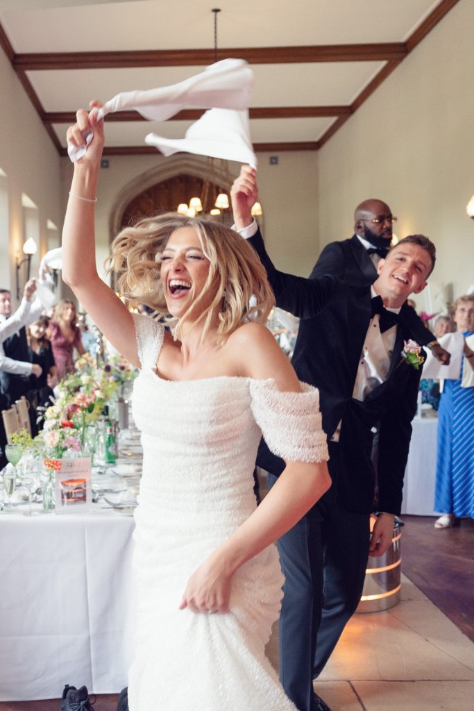 A joyful bride in a white dress and a groom in a tuxedo dance and wave serviettes in the air at a lively wedding reception, surrounded by smiling guests in a decorated hall.