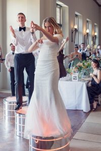 A bride in a white dress and a groom in a tuxedo joyfully dance on glowing circular platforms at a wedding reception, surrounded by smiling guests seated at decorated tables.