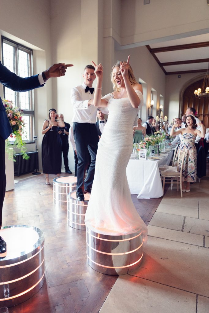A bride in a white dress and a groom in a tuxedo dance joyfully on glowing round platforms at a wedding reception, surrounded by smiling guests in a bright, elegant hall.