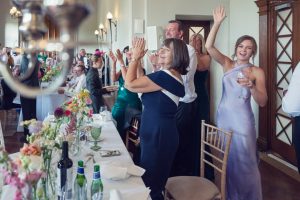 A group of well-dressed guests stand and cheer at a decorated banquet table during a celebration, smiling and raising their hands in excitement. Flowers, glasses, and bottles are visible on the table.