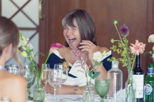 A woman with shoulder-length grey hair laughs joyfully at a table decorated with colourful flowers, glassware, and bottles. She wears an off-the-shoulder dress and is engaged in conversation with someone out of view.