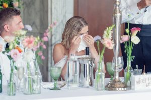 A bride wipes tears from her eyes with a tissue whilst sitting at a decorated table next to the groom during a wedding reception. Glasses, flowers, and a cake are visible on the table.