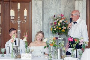 A man in a dinner jacket speaks into a microphone at a wedding reception table. A smiling bride and groom sit beside him surrounded by flowers, candles, and glassware, with a marble wall and large floral arrangement in the background.