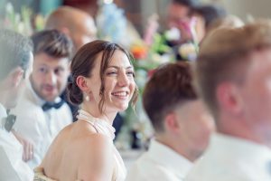 A woman in a light dress smiles brightly whilst seated at a table with several people, all dressed formally. The background is softly blurred, with colourful flowers visible.