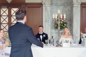 A man in a dinner jacket stands facing a long table where a bride, groom, and guests sit; the bride looks surprised and delighted. The setting features elegant decor, a candelabrum, and floral arrangements.