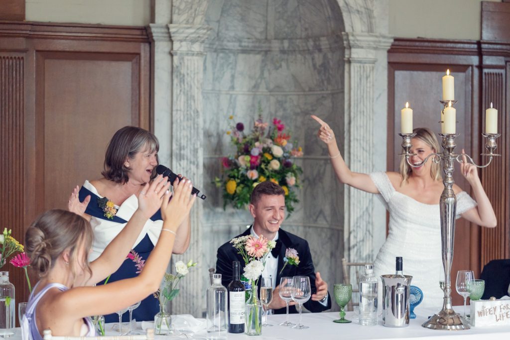 A bride, groom, and guests at a wedding reception laugh and celebrate at a table with flowers, wine, and candles. The bride and a woman beside her raise their arms joyfully, creating a lively scene.