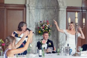 A bride, groom, and guests at a wedding reception laugh and celebrate at a table with flowers, wine, and candles. The bride and a woman beside her raise their arms joyfully, creating a lively scene.