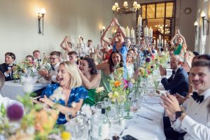 A joyful crowd at a formal event sits at long tables decorated with flowers and candles, clapping and cheering. Many guests are smiling, some with hands raised in celebration, creating a lively, festive atmosphere.