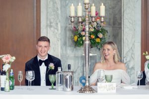 A bride and groom sit smiling at a reception table decorated with flowers, wine glasses, and a candelabrum. The bride wears an off-the-shoulder dress and the groom wears a dinner suit. A Wifey for Lifey sign is on the table.