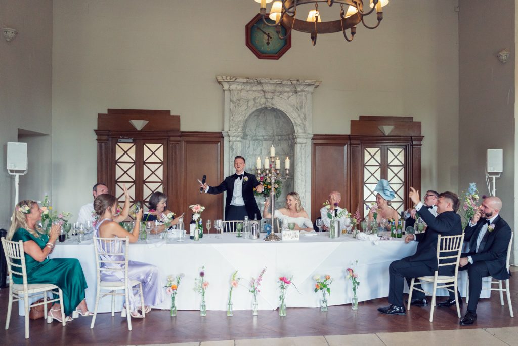 A wedding reception: a man stands and gives a toast at a long, flower-decorated table, with guests on both sides raising glasses and smiling in a bright, elegant room with a marble fireplace in the background.