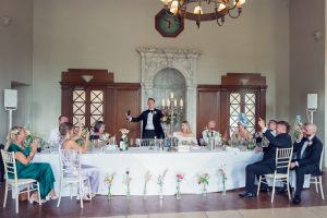 A wedding reception: a man stands and gives a toast at a long, flower-decorated table, with guests on both sides raising glasses and smiling in a bright, elegant room with a marble fireplace in the background.
