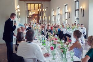 A man in a dinner jacket gives a speech with a microphone at a long, elegant wedding reception table as guests listen and raise glasses in a bright, ornate room with flowers and candelabra.