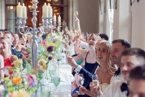 A group of people dressed formally sit at a long, decorated table, raising glasses for a toast at a celebration or wedding, with flowers and candle holders as centrepieces. Everyone appears happy and festive.