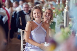 A young woman in a light purple dress sits on a white chair, smiling and laughing at a lively event. Other guests, including a woman behind her, are also smiling and clapping amid colourful decorations.