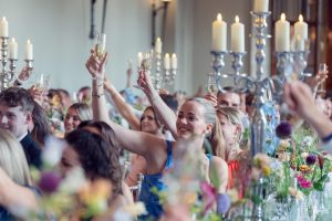 A group of people dressed formally raise glasses in a toast at a decorated event with floral arrangements and tall candlesticks, smiling and celebrating together in a bright, elegant setting.