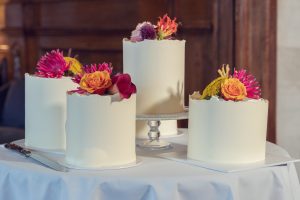 Four white frosted cakes, each topped with colourful flowers, are arranged on a round table covered with a white tablecloth. One cake is elevated on a glass stand. A cake knife is placed nearby.