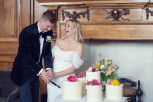 A bride and groom laugh joyfully whilst cutting a wedding cake together. They stand in front of a wooden fireplace, with several white cakes decorated with colourful flowers on a table before them.