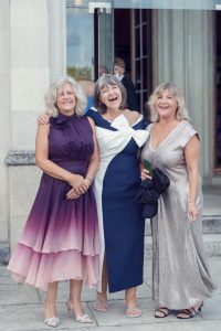 Three smiling women in elegant dresses stand arm-in-arm outdoors in front of a building entrance, posing happily for a photo.
