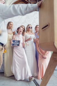 Six women in pastel dresses pose for a photo at a wedding. One holds a Best Wedding Ever sign, and another wears white sunglasses. A man's arm and a photo booth are visible in the foreground.