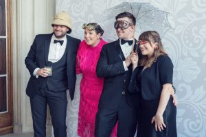 Four people dressed in formal attire pose playfully together. Two men wear dinner jackets, with one in a pith helmet and the other holding a parasol and wearing goggles. Two women wear bright accessories, with one in a vibrant pink dress.