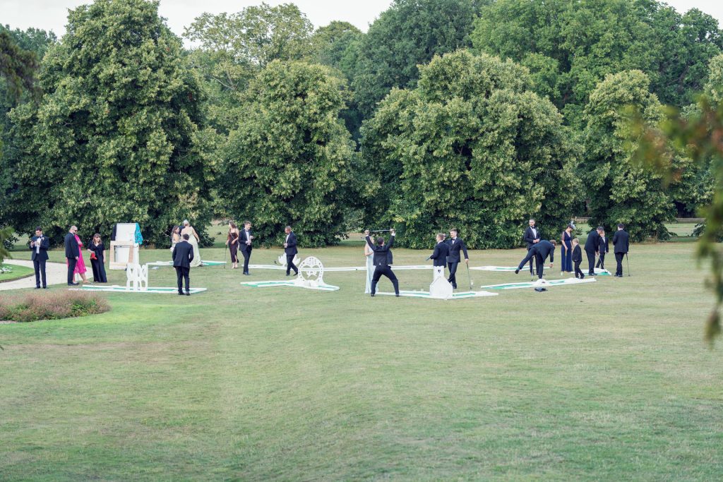 A group of people dressed mostly in black are gathered outdoors on a grassy lawn, interacting with large white sculptures and objects, surrounded by tall green trees.