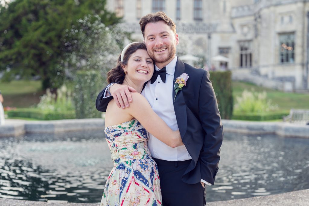 A smiling couple pose in front of a fountain, dressed in formal attire; the woman wears a colourful strapless dress and the man wears a black tuxedo with a buttonhole. A large building and greenery are in the background.