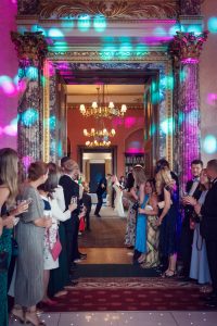 A bride and groom share a kiss at the end of a grand corridor, surrounded by guests on both sides. Colourful lights add a festive atmosphere in the ornate venue. Guests watch and celebrate, some holding phones to capture the moment.