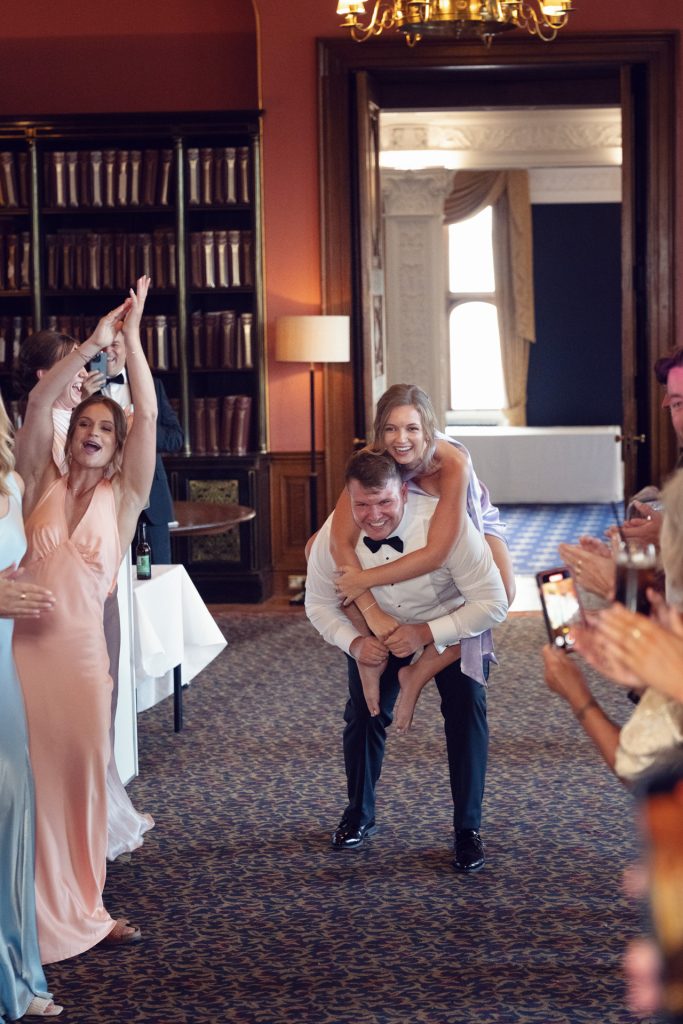 A smiling man in a tuxedo gives a piggyback ride to a laughing woman in a lavender dress as people around them clap and cheer in a formal, wood-panelled room with bookshelves.