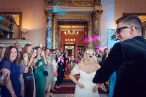 A bride and groom in sunglasses dance joyfully at their wedding reception, surrounded by smiling and cheering guests in a grand, elegant room with ornate columns and chandeliers.