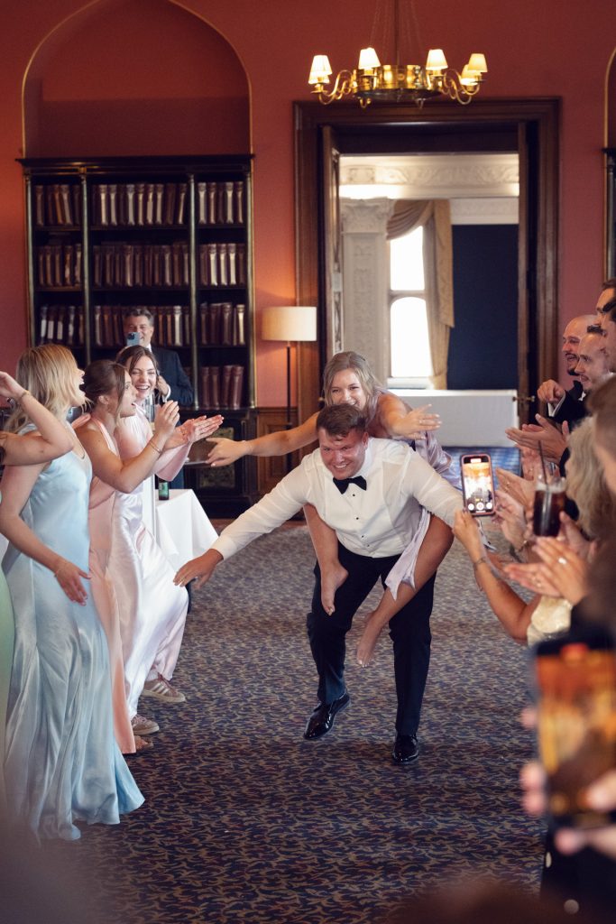 A smiling man in formalwear gives a piggyback ride to a laughing woman in a white dress as guests in pastel gowns and suits cheer and clap in an elegant, warmly lit room with bookshelves and arched doorways.