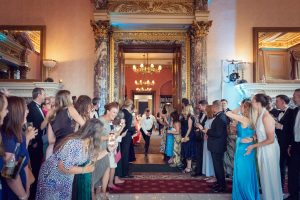A crowd in formal attire lines a grand hallway, cheering and taking photos as a couple, possibly just married, walks in from double doors in an elegant, chandelier-lit venue.