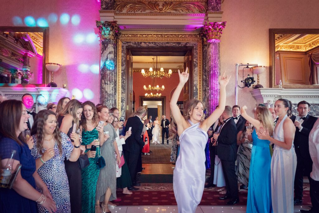 A woman in a satin dress dances with her arms raised at a formal event, while a crowd of well-dressed guests smiles and watches her in an elegant, ornate room decorated with columns and chandeliers.