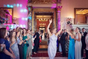 A woman in a satin dress dances with her arms raised at a formal event, while a crowd of well-dressed guests smiles and watches her in an elegant, ornate room decorated with columns and chandeliers.