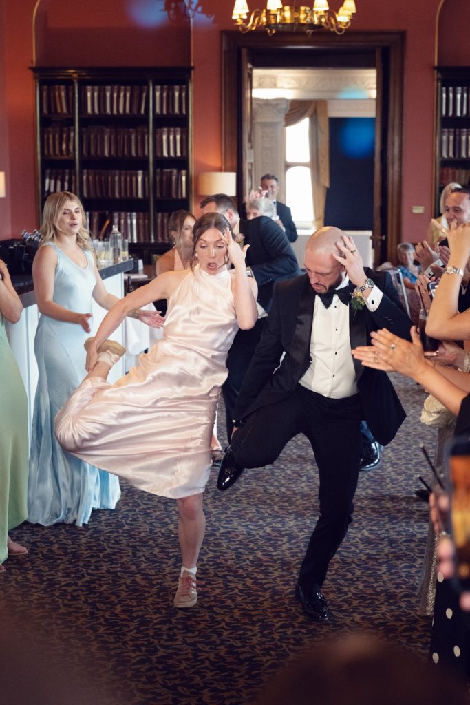 A woman in a pale pink dress and a man in a tuxedo dance energetically together at an indoor event, surrounded by cheering guests in formal attire.