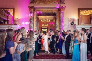A bride and groom dance joyfully in the centre of an elegant ballroom, surrounded by guests dressed in formal attire who are smiling, clapping, and capturing the moment with their phones.
