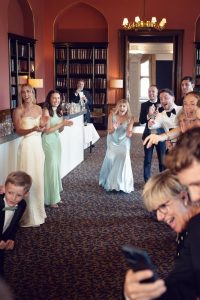 A group of well-dressed people at a formal event stand in a room with bookshelves, clapping and smiling excitedly towards the camera. The atmosphere is joyful and celebratory.