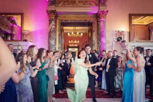 A joyful crowd in formal attire gathers around a dancing couple at an opulent indoor venue with marble columns and chandeliers, applauding and smiling during a celebration.
