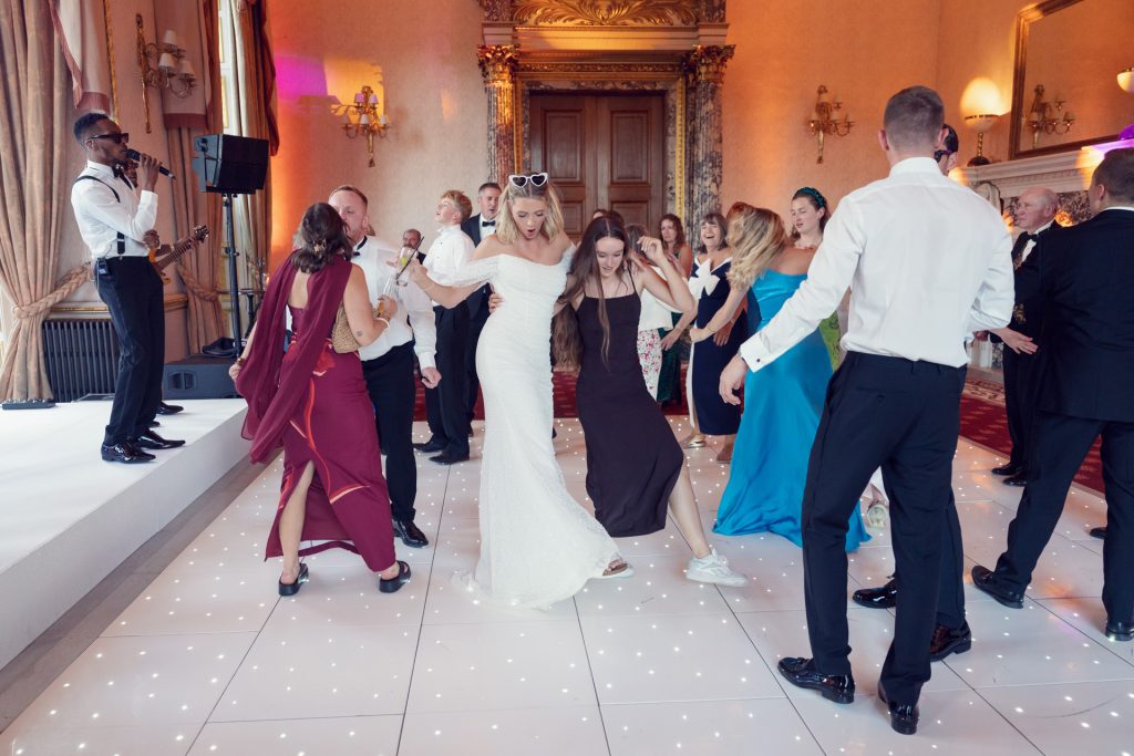 A group of people dressed formally dance and celebrate on a lit-up floor in an elegant, ornate room, whilst a singer and band perform on stage in the background.