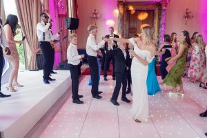 A lively group of people, including children and adults in formal attire, dance on a lit-up floor at an elegant indoor event, whilst a live band performs on stage. Pink and purple lights illuminate the ornate room.