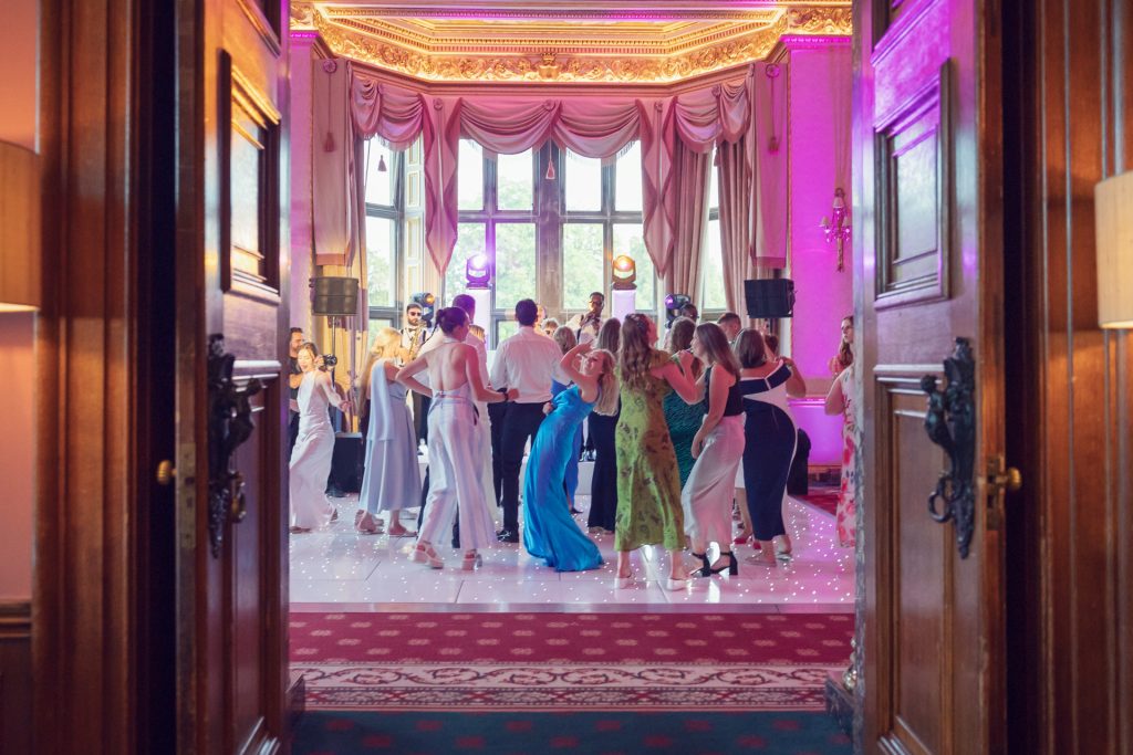 A group of people dressed in formal wear dance on a lighted floor in an elegant, pink-lit room with tall windows, seen through open double doors.